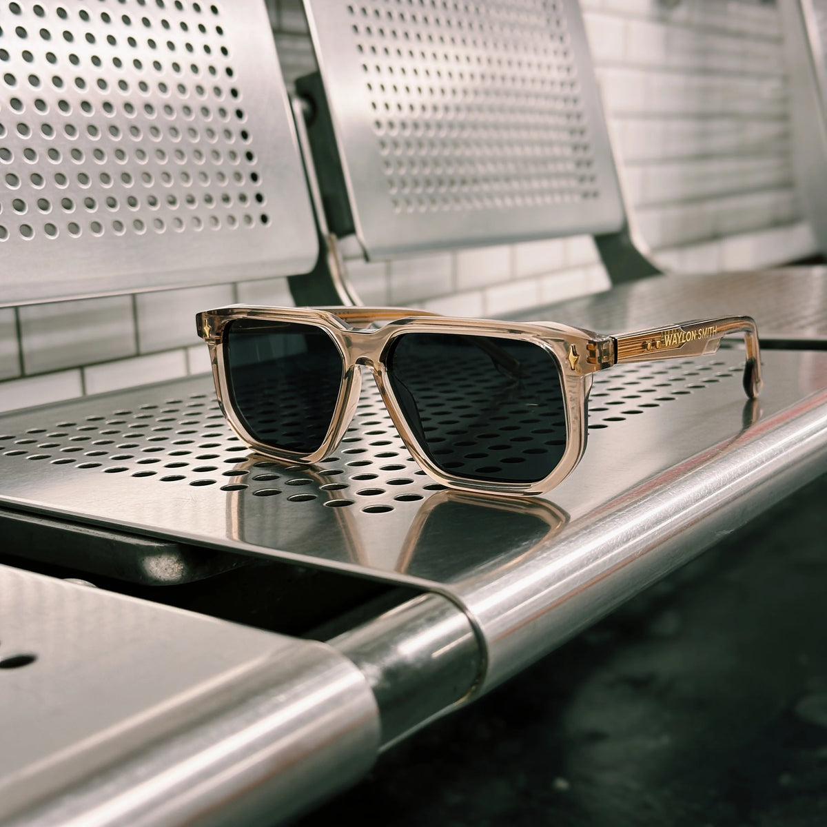 Transparent champagne Waylon Smith sunglasses resting on a metal subway bench