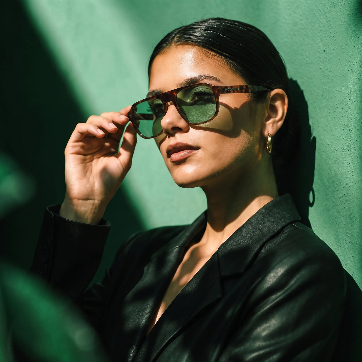 Woman in a leather jacket adjusting tortoiseshell Waylon Smith sunglasses with green lenses against a green wall