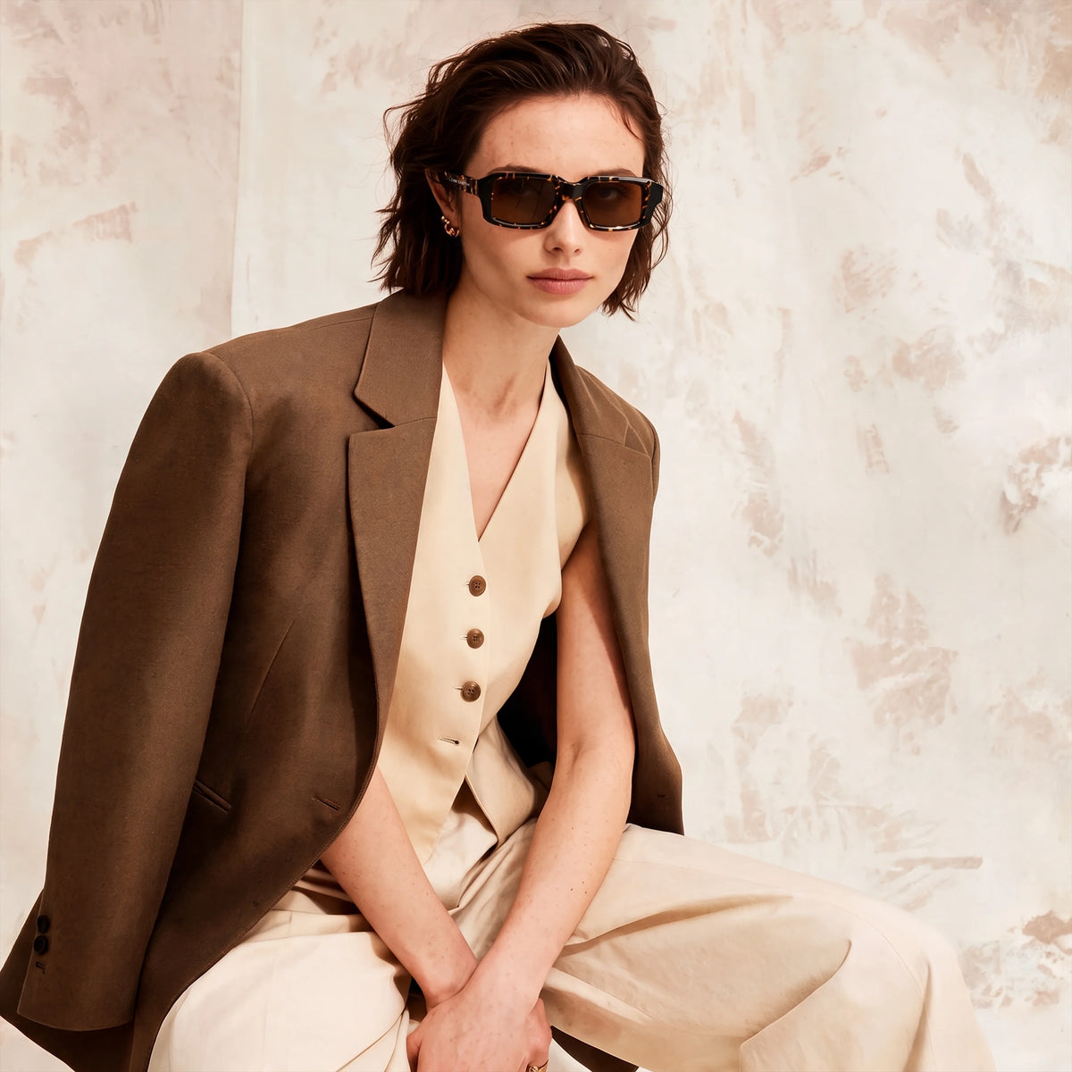 Woman in a brown suit wearing tortoiseshell Waylon Smith sunglasses, seated against a textured backdrop