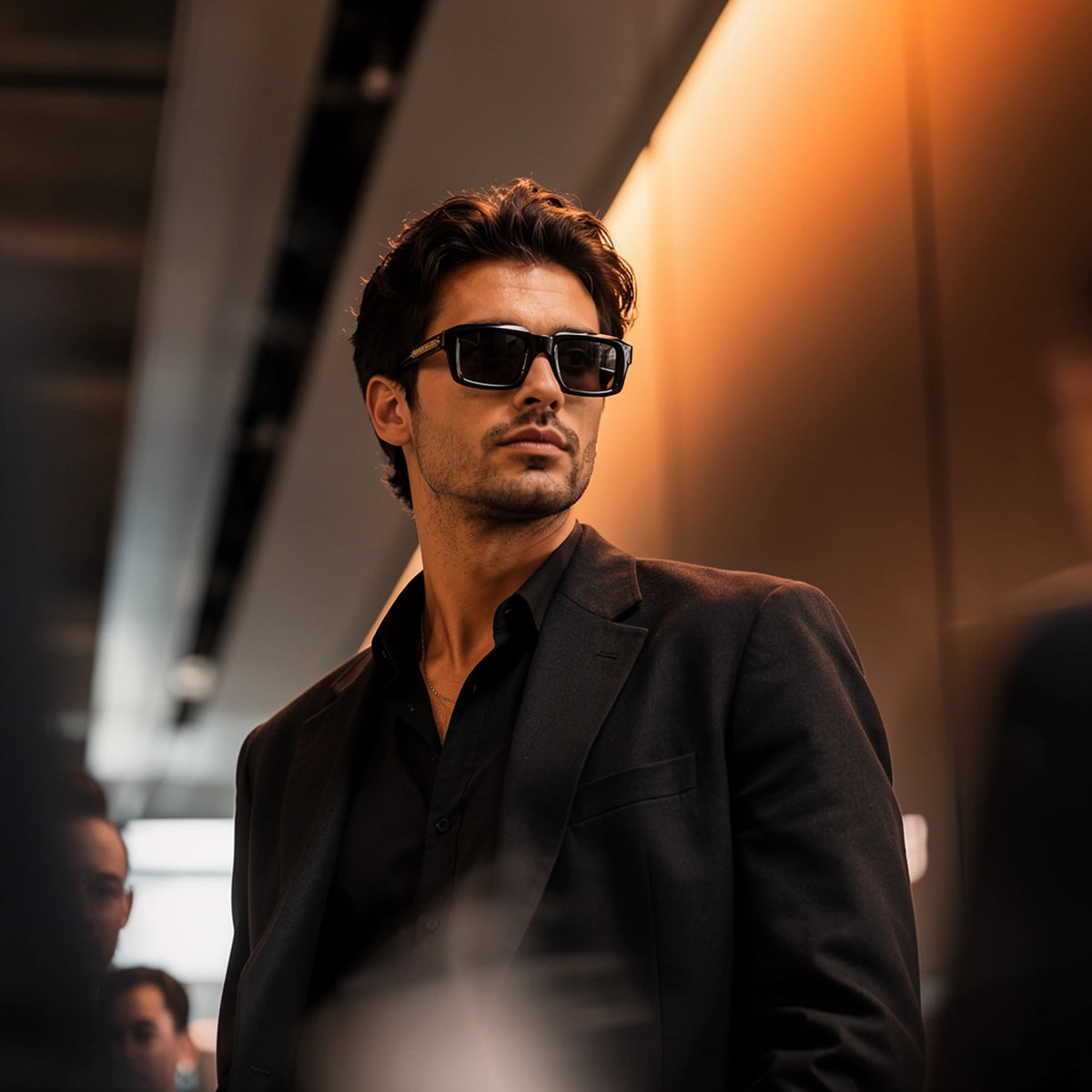 Man in a black suit wearing black Waylon Smith sunglasses, standing in a warmly lit train