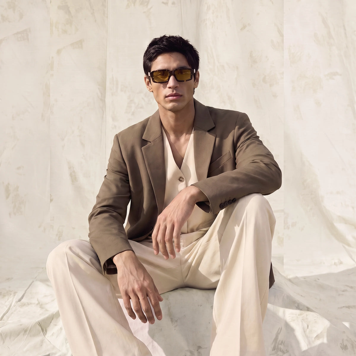 Man in a beige suit wearing tortoiseshell Waylon Smith sunglasses, seated against a draped fabric backdrop