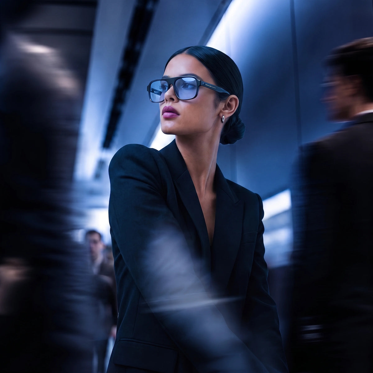 Woman in a black suit wearing black Waylon Smith sunglasses with blue lenses in a dimly lit corridor