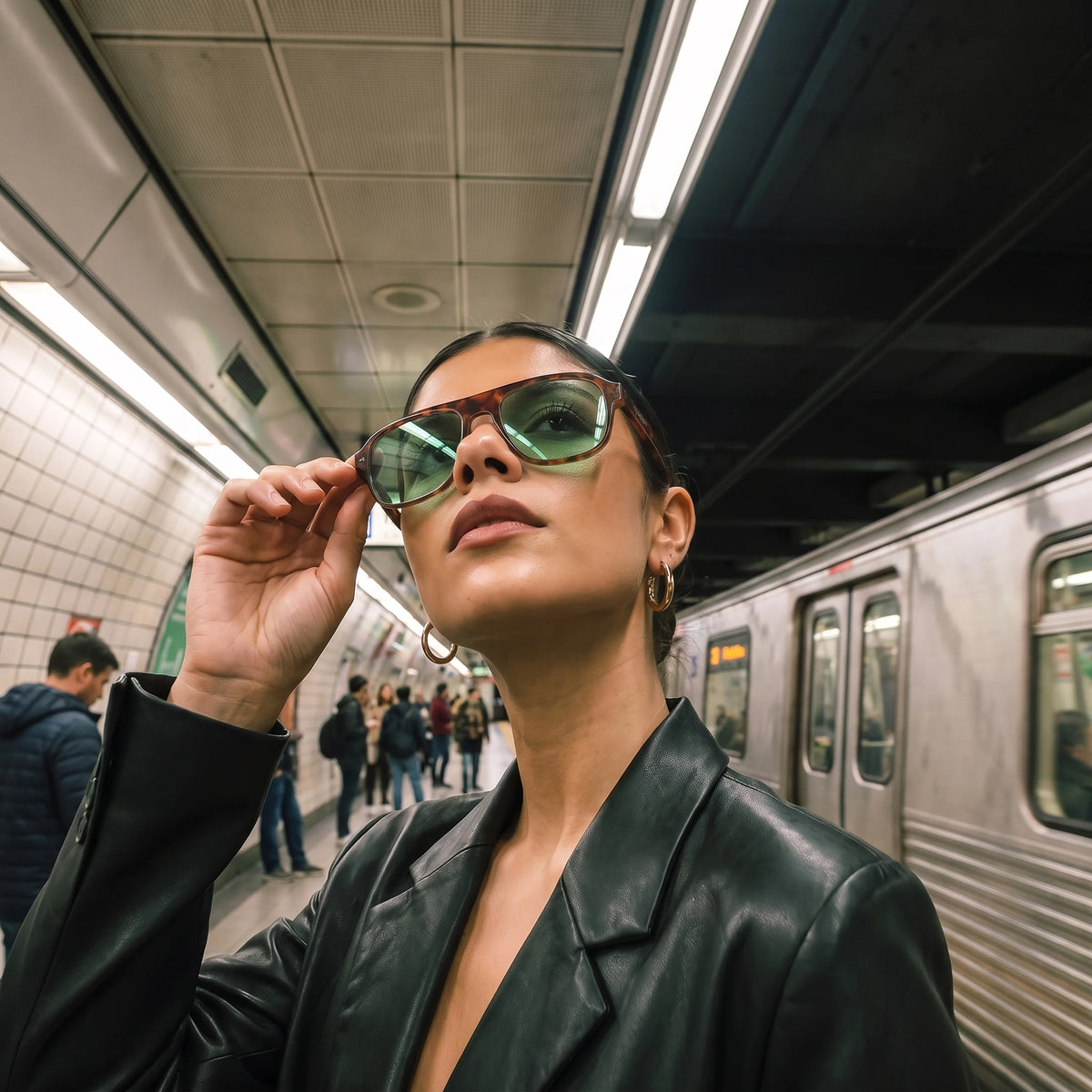 Woman in a leather jacket adjusting tortoiseshell Waylon Smith sunglasses with green lenses on a subway platform