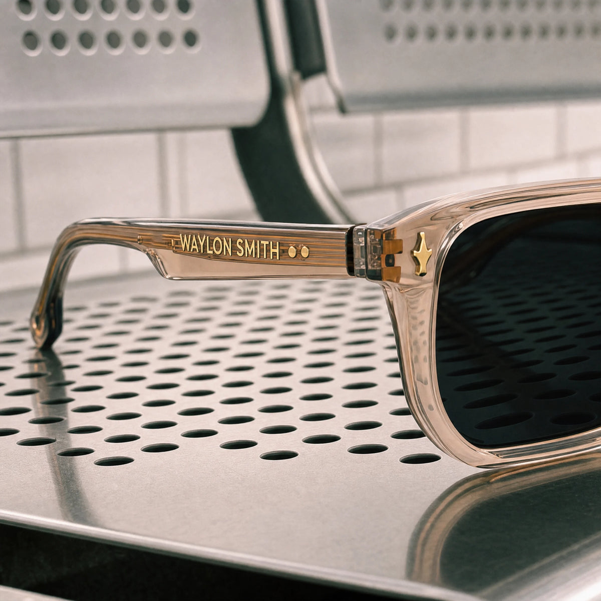 Close-up of transparent champagne Waylon Smith sunglasses on a perforated metal surface, showing the Waylon Smith wordmark on the temple