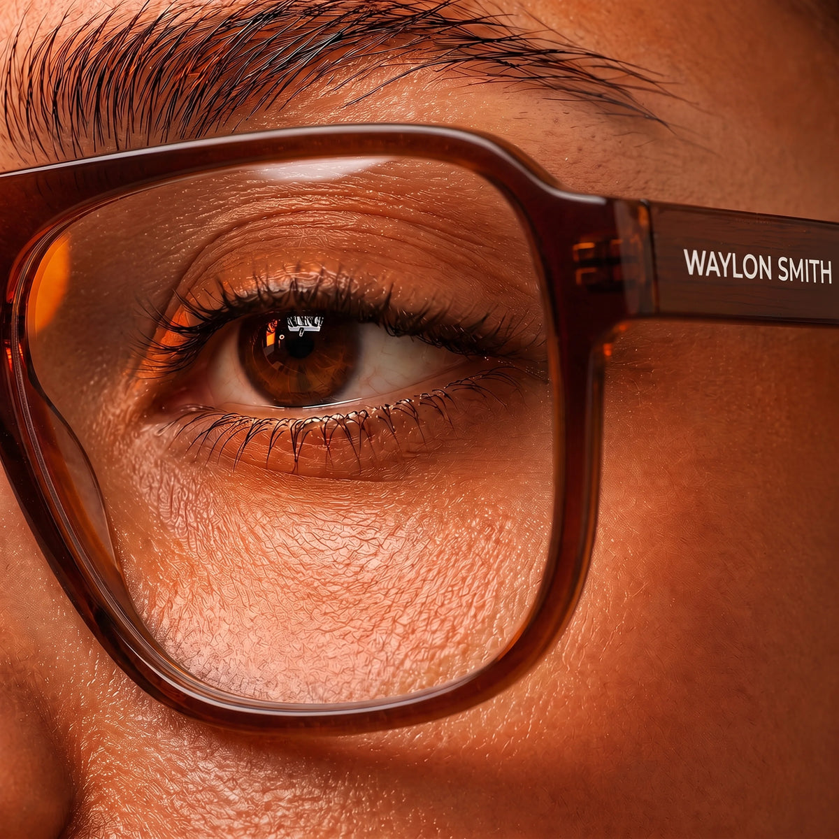 Extreme close-up of an eye through amber tortoiseshell Waylon Smith sunglasses, showing the Waylon Smith wordmark on the temple