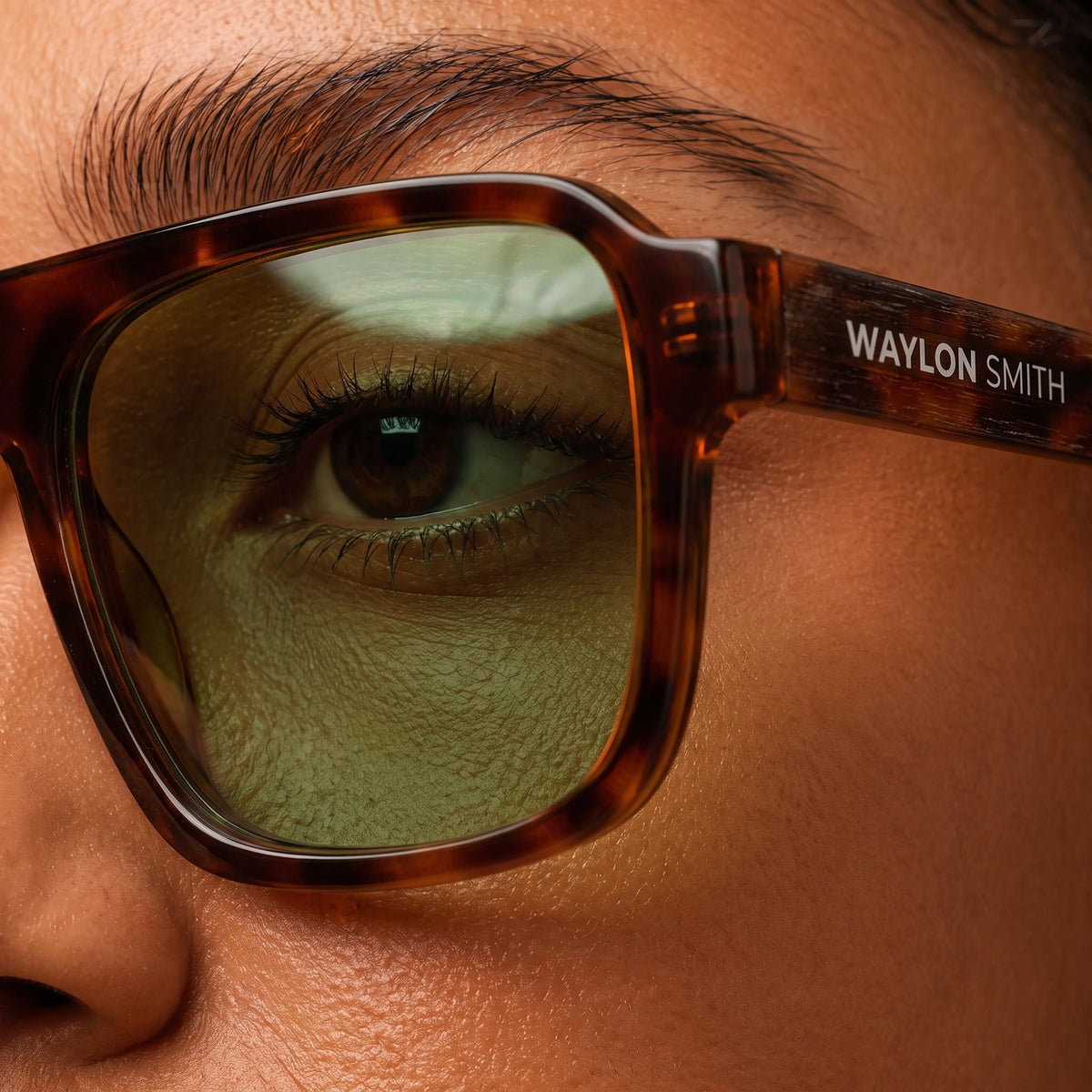 Extreme close-up of an eye through tortoiseshell Waylon Smith sunglasses with green lenses, showing the Waylon Smith wordmark on the temple