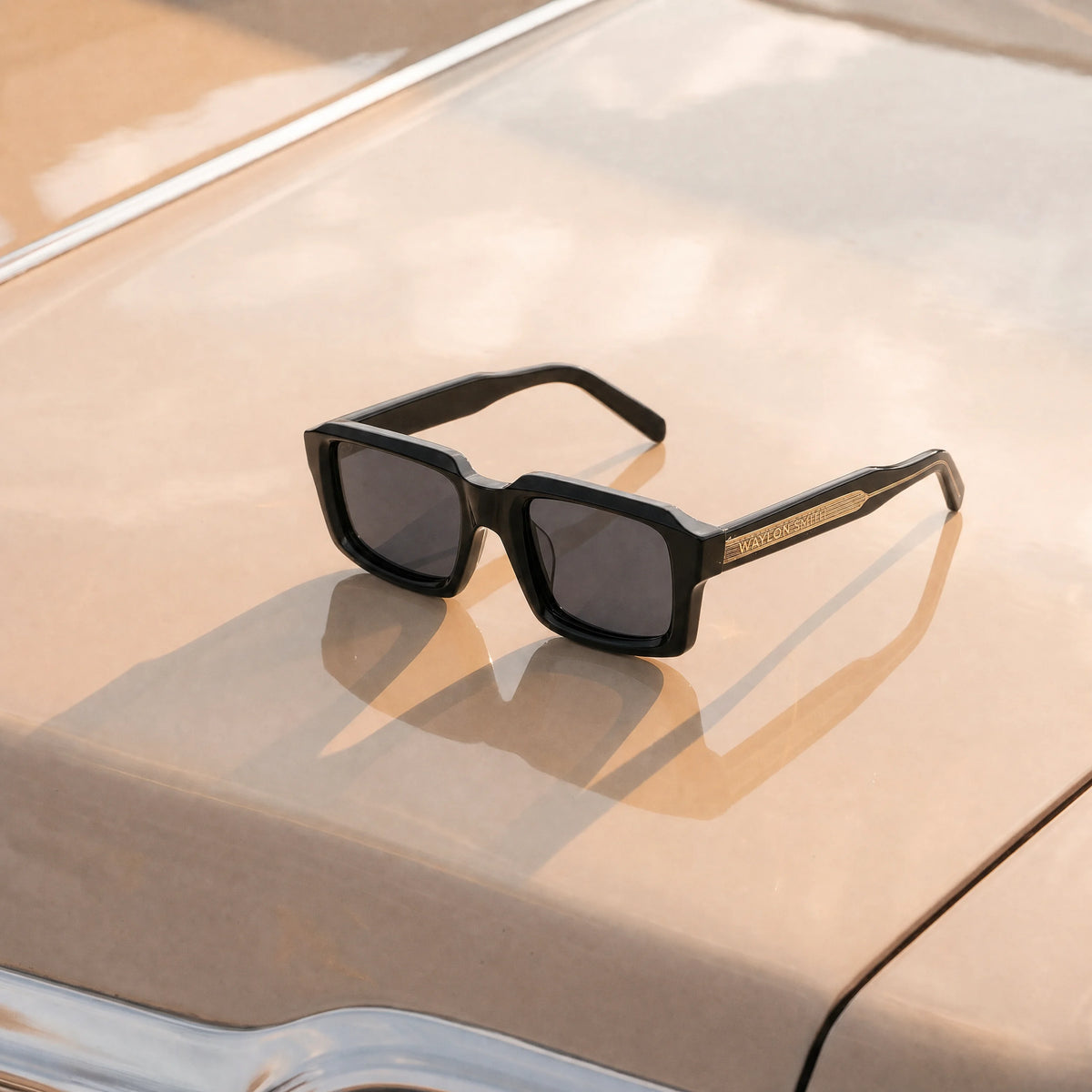 Black Waylon Smith sunglasses with a gold textured temple, resting on the hood of a car