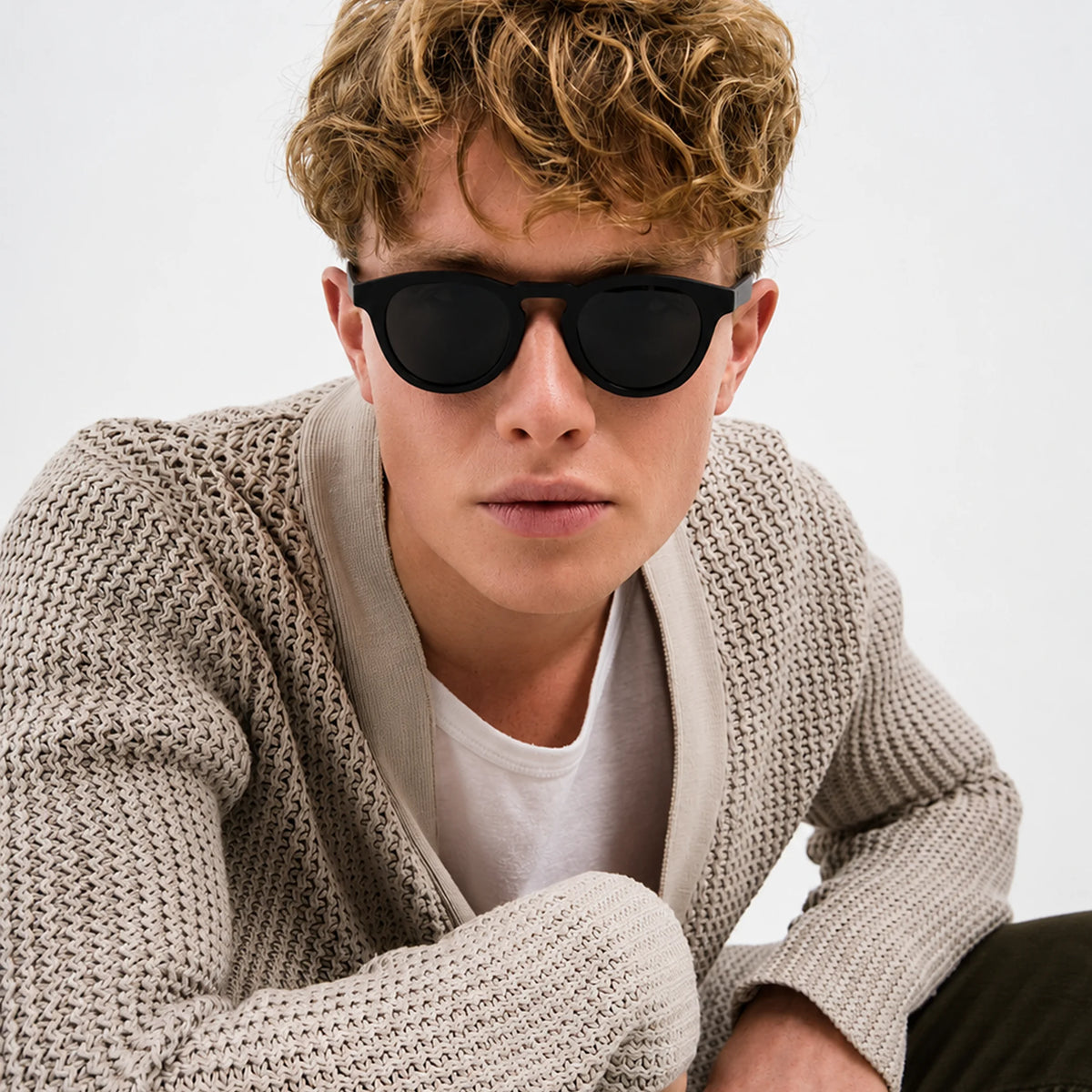 Man with curly hair wearing round black Waylon Smith Midnight Havana sunglasses and a cream knit cardigan